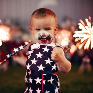 An image capturing the joyous spirit of a toddler boy on the 4th of July, dressed in a patriotic outfit adorned with stars and stripes, holding a miniature American flag while surrounded by festive fireworks